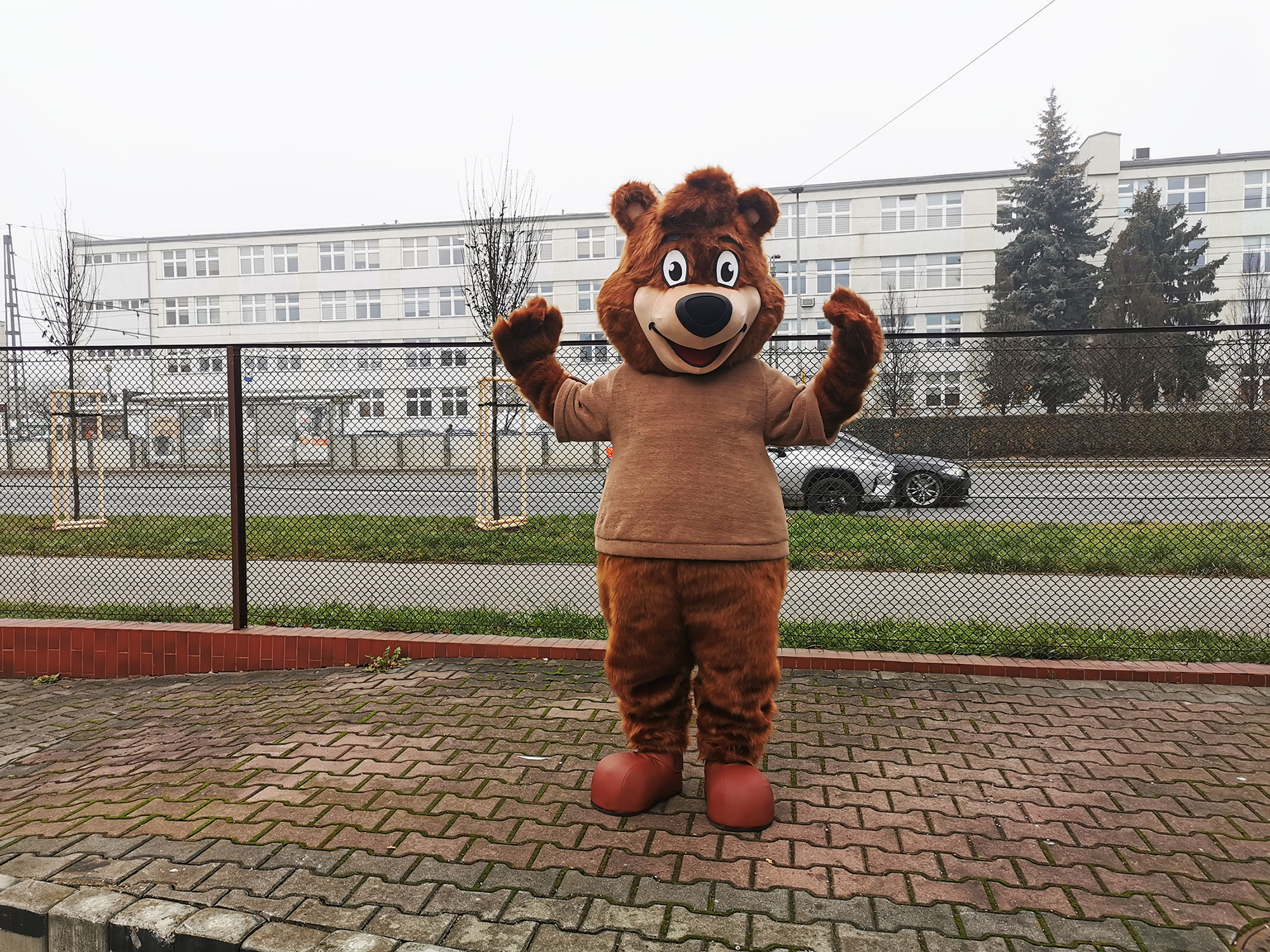 Na fotografii widnieje kostium reklamowy stylizowany na misia w swetrze, pozujący do zdjęcia przed siedzibą firmy Elabika